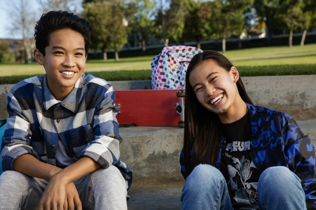 Two smiling teenagers sitting on a concrete ledge, showcasing their confidence and camaraderie, with a colorful backpack and skateboard in the background, representing the joy of youth and social interactions.